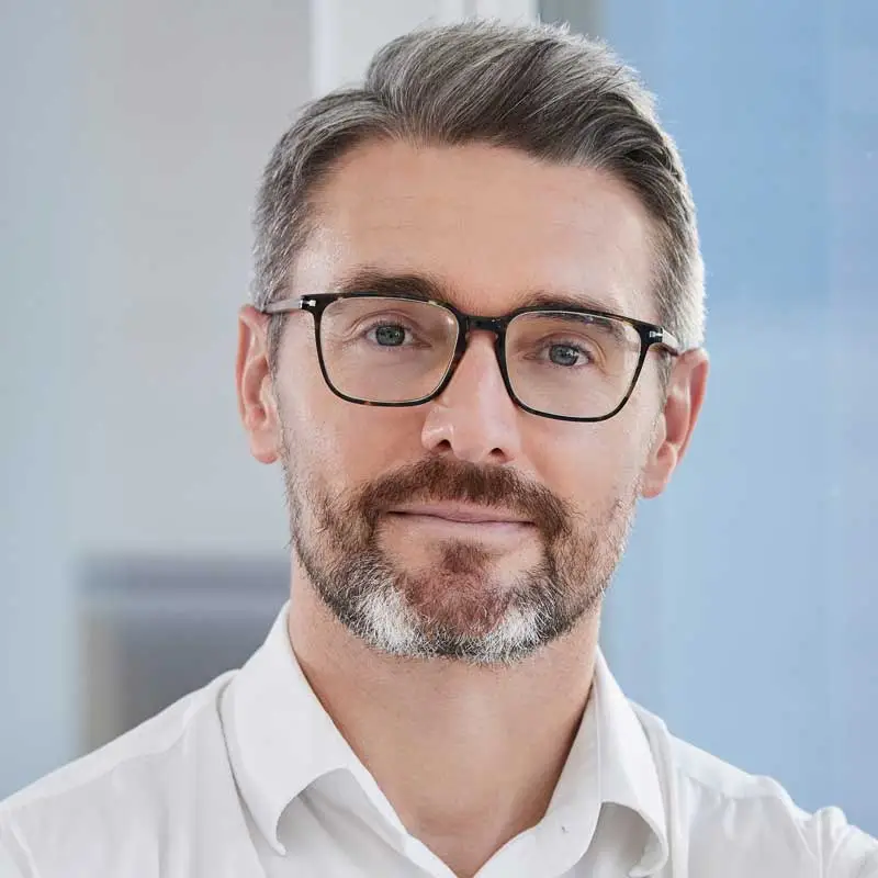 Portræt af mand med gråligt hår og skæg, iført briller og hvid skjorte.